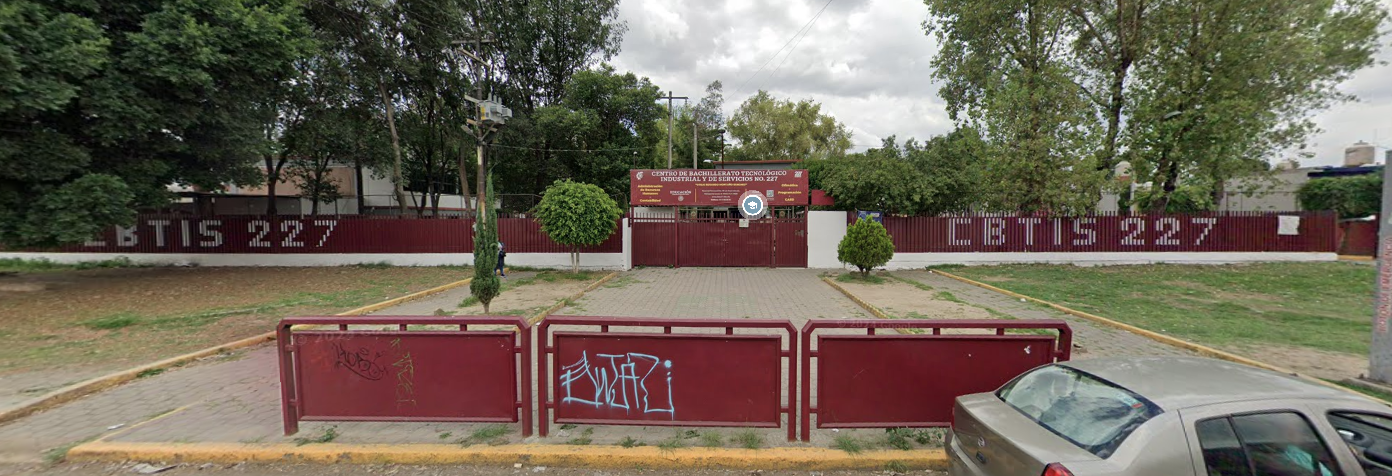 Fotografía en la que se muestra la entrada del plantel, conformada por rejas de color rojo que rodean las instalaciones. Sobre las rejas están pintadas letras blancas que dicen Cebetis 227. En medio de las rejas hay un portón rojo que da acceso al plantel.
